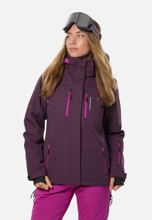 Femme portant une veste de ski violette avec des fermetures éclair roses et un pantalon rose assorti, regardant légèrement sur le côté, avec des lunettes de ski sur le front.