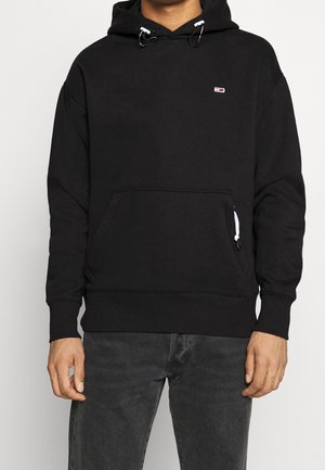 Homme portant un sweat à capuche noir avec un petit logo rectangulaire sur la poitrine, une poche à l'avant et des cordons blancs, associé à un jean foncé.