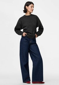 Jaunas žmogus, vilkintis laisvu juodu megztiniu, plačių juodų džinsų ir raudonais batais, stovintis su rankomis kišenėse ant baltos fono.