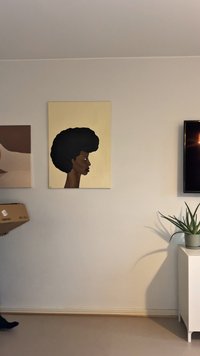 Stranska profilna slika osebe z afro pričesko na bež steni, na desni televizija, rastlina v lončku na belem omarici in oseba, ki nosi škatlo.