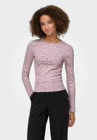 Różowa bluzka z długim rękawem w prążki z białym kwiatowym wzorem, z okrągłym dekoltem i falbanką na dole, zestawiona z czarnymi spodniami.