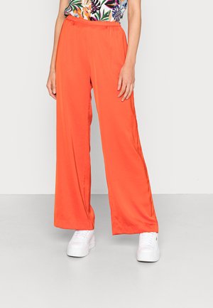 Calças largas laranja com cintura elástica, feitas de um tecido leve. Combinadas com ténis brancos para um look casual.
