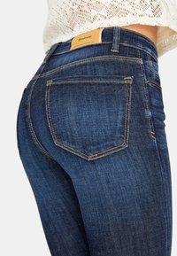 Jeans de mezclilla azul oscuro con un diseño ajustado, que presentan dos bolsillos traseros y costuras doradas de contraste en los bordes. Etiqueta de marca de cuero visible.