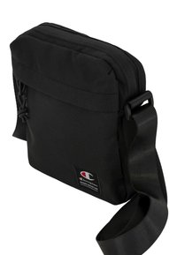 Borsa a tracolla nera in poliestere con chiusura a zip, tracolla regolabile e un piccolo dettaglio del logo. Superficie liscia e forma rettangolare.