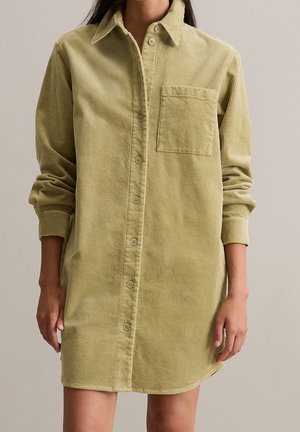 Vestido camisa de pana beige de manga larga con botones en la parte delantera y un bolsillo en el pecho, usado con las mangas remangadas.