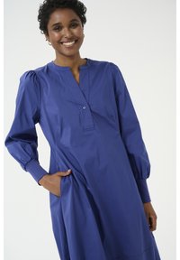 Karaliski zils kleita, kas izgatavota no viegla auduma. Aprīkota ar pogu slēgšanu, garām pūkainām piedurknēm un sānu kabatām praktiskai lietošanai.