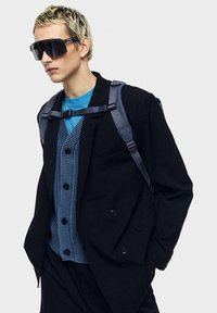 Svart blazer över en blå ribbad väst, bärs med en blå t-shirt. Utrustad med stora svarta solglasögon och en grå ryggsäck med justerbara remmar.