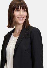 Blazer nero su misura con vestibilità slim, rever a punta e una tasca sul petto, abbinato a un top crema plissettato. Tessuto dalla consistenza morbida.