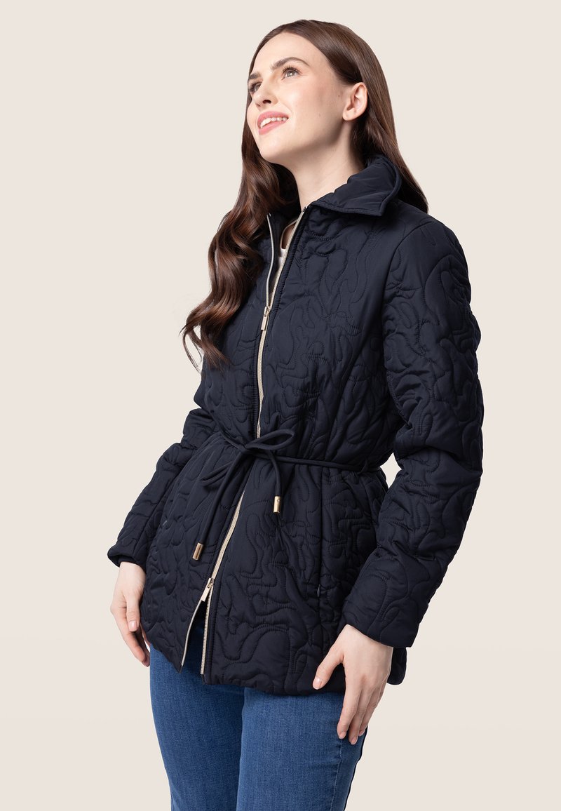 Veste matelassée noire avec fermeture éclair dorée et taille à cordon, dotée d'un col montant et d'un motif texturé. Portée sur un jean bleu.