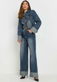 Denimjacka och vida jeans-set, i mellanblå färg, med kontrasterande manschetter, knappstängning och framfickor. Slät textur.