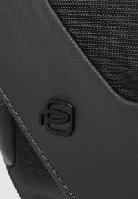 Bolso negro texturizado con un acabado suave, que presenta un logo sutil, costuras contrastantes y un elemento de diseño moderno.