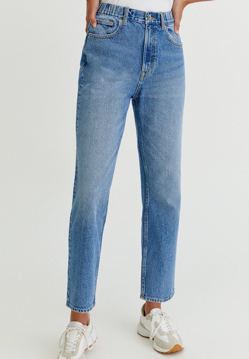 Personne portant un jean bleu taille haute à jambes droites et des baskets blanches, debout les mains dans les poches sur un fond uni.
