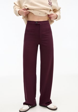 Mujer levantando un jersey crema para mostrar unos pantalones burdeos de cintura alta y pernera ancha combinados con zapatillas blancas y beige.