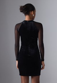 Robe en velours noir avec de longues manches en tissu transparent, coupe ajustée, col montant et détail de fermeture éclair dans le dos, se terminant au-dessus du genou.