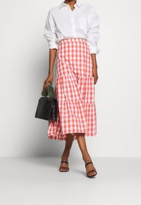 Rote und weiße Karoschürze mit gestuften Rüschen, kombiniert mit einem weißen Hemd und einer schwarzen Handtasche. Flache schwarze Sandalen runden das Outfit ab.