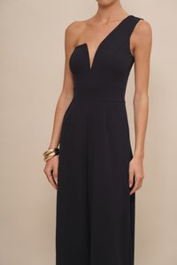Marineblauwe jumpsuit met één schouder, diepe V-hals, flatterende taille naad en wijde pijpen; stof heeft een gladde textuur. Geaccentueerd met gouden armbanden.