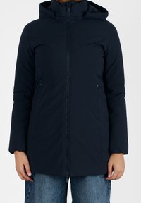 Femme portant un manteau d'hiver à capuche zippé bleu marine foncé avec des poches latérales et un jean bleu, se tenant contre un fond blanc.