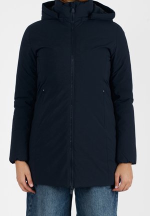 Kvinde iført en mørkeblå lynlås vinterjakke med hætte og sidelommer samt blå jeans, stående foran en hvid baggrund.