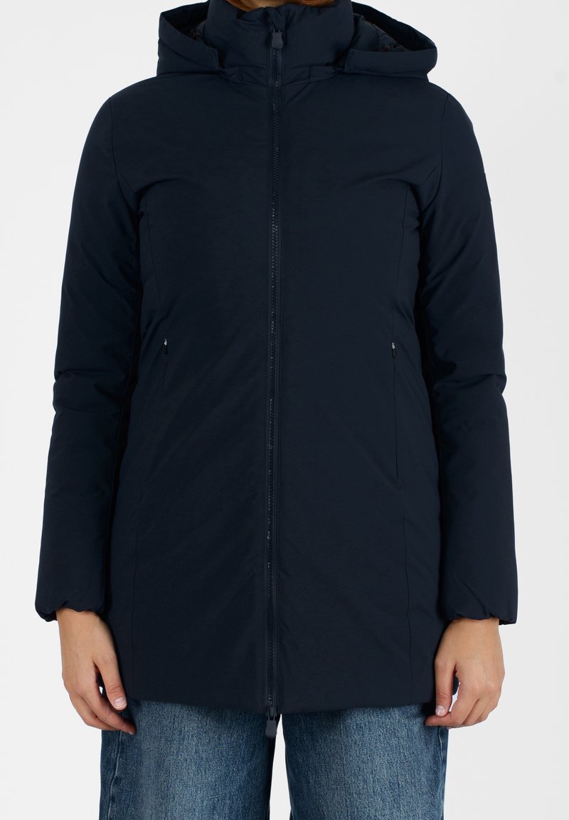 Femme portant un manteau d'hiver à capuche zippé bleu marine foncé avec des poches latérales et un jean bleu, se tenant contre un fond blanc.