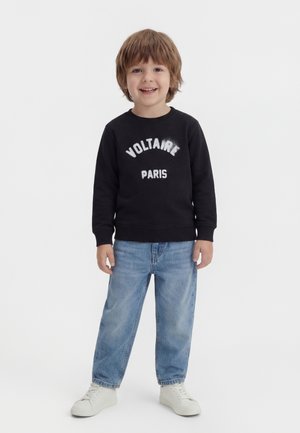 Χαμογελαστός νεαρός αγόρι με μεσαίο καστανό μαλλί που φοράει μαύρο φούτερ με το κείμενο "Voltaire Paris", ανοιχτό μπλε τζιν και λευκά αθλητικά παπούτσια.