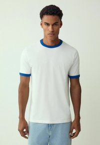 T-shirt bianco a maniche corte con colletto e polsini in maglia a contrasto blu, realizzato in morbido cotone. Il modello indossa jeans azzurri chiari.