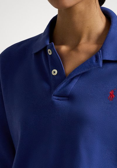 Modra polo majica iz teksturirane tkanine z ovratnikom, opremljena s tremi gumbi in majhnim rdečim vezenim logotipom na levi strani prsnega koša.