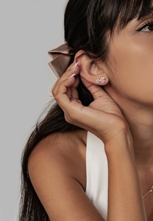 Frau mit diamantenförmigen Ohrringen in Blumenform, die ein Ohrläppchen mit manikürten Fingern berührt, das Haar mit einem beigen Band zurückgebunden.