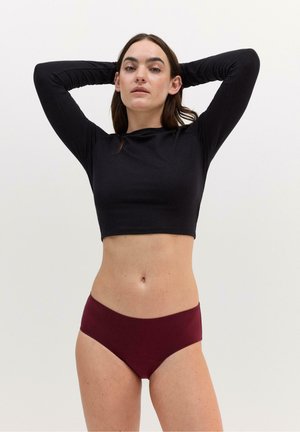 Kvinde står med hænderne bag hovedet iført en sort langærmet crop top og bordeaux undertøj mod en ensfarvet baggrund.