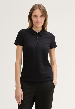 Jeune femme aux cheveux châtain clair et lisses, portant un polo noir à manches courtes et un pantalon noir, debout devant un fond uni.