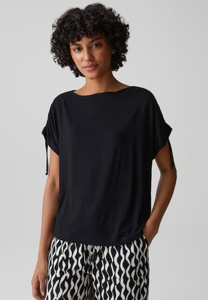 Donna con capelli ricci che indossa una maglietta nera a maniche corte e pantaloni con motivo ondulato bianco e nero, in piedi davanti a uno sfondo semplice.