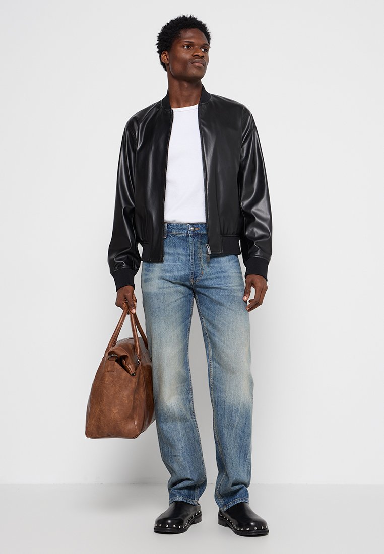 Veste bomber en simili cuir noir, t-shirt blanc, jeans déchirés bleu clair, sac fourre-tout en cuir marron et sabots noirs avec des clous argentés.