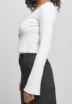 Kvinde vist fra siden iført en tætsiddende hvid langærmet crop top med udvidede manchetter og sort højtaljet denimjeans.