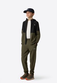 Παιδικό αθλητικό σύνολο που περιλαμβάνει μαύρο και ελιά φερμουάρ μπουφάν, παντελόνια σετ, και γκρι-πορτοκαλί αθλητικά παπούτσια, συμπληρωμένο με ένα ναυτικό καπέλο.