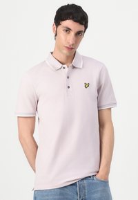 Polo shirt lavande clair en tissu doux. Il présente un col classique, une patte de boutonnage à trois boutons et un petit logo de papillon jaune brodé.
