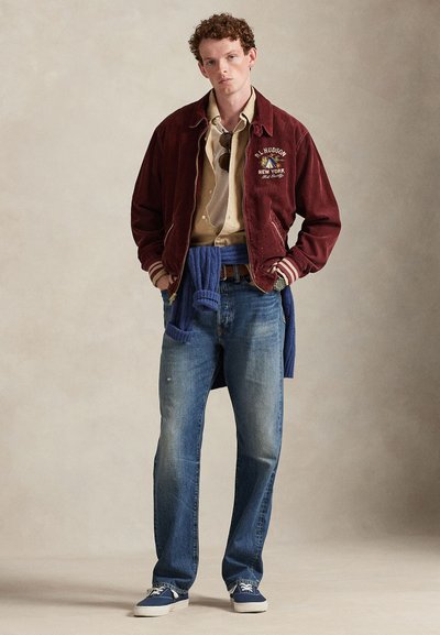 Bordeauxrood corduroy jasje met witte strepen, beige overhemd, blauwe spijkerbroek en marineblauwe schoenen. Opvallende details zijn onder meer logo borduursel en een trui die om de taille is geknoopt.