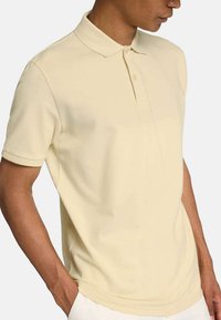 Polo shirt jaune pâle avec col, manches courtes et patte de boutonnage à deux boutons. Texture lisse avec une silhouette ajustée. Pas de motifs visibles.