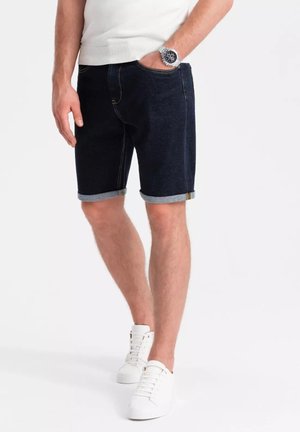 Jeansshorts, dunkelblau mit einem ausgebleichten Finish. Umschlagbündchen, Fünf-Taschen-Design und goldene Nähte. Kombiniert mit weißen Sneakers.
