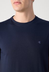 Tricou din bumbac de culoare bleumarin, cu mâneci scurte, decolteu rotund și un mic logo brodat pe partea stângă a pieptului. Textură netedă și ajustată.
