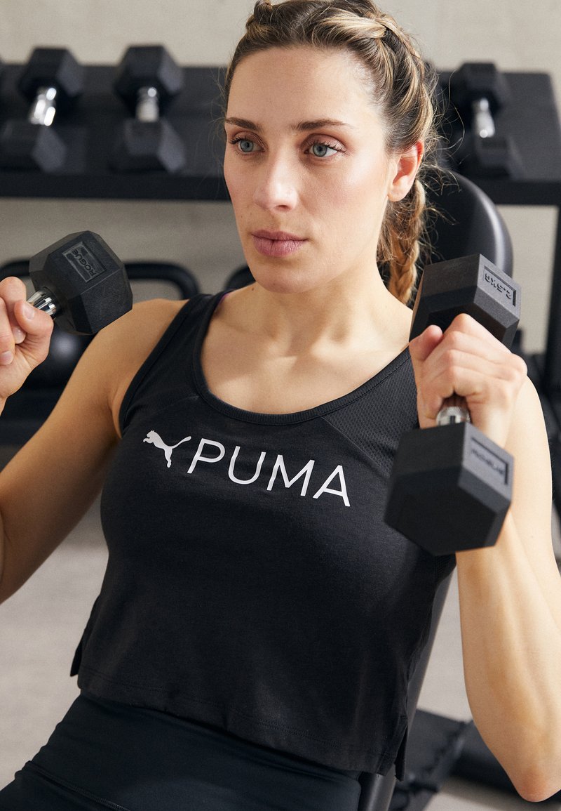 Fekete, rövidítő tank top, fehér "PUMA" logóval. Egyén fekete, hatszögletű súlyzókat tart. A háttérben elmosódott súlyzósor látható.
