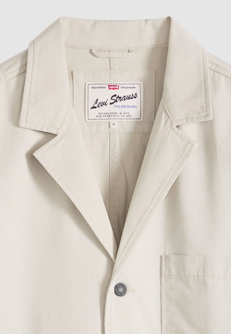 Veste beige Levi Strauss avec col cranté, un bouton, poche poitrine, et étiquette Levi's à l'intérieur du col indiquant la marque et la taille moyenne.