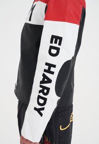 Svart, röd och vit läderjacka med en framträdande "ED HARDY"-logotyp på ärmen, kontrasterande texturer och en strukturerad design.