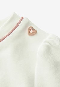 Bílá látka s texturovaným povrchem, zdobená růžově zlatou sponou ve tvaru srdce s květinovými detaily a růžovým akcentem podél výstřihu.