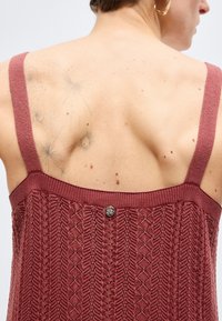 Γυναίκα που φοράει αμάνικη υφασμάτινη μπορντό μπλούζα, η οποία δείχνει ένα αχνό τατουάζ από σπόρους πικραλίδας στην πάνω αριστερή πλάτη της και μικρές σκούρες ελιές στο δέρμα της.