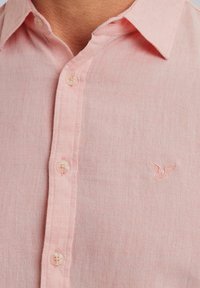 Camisa de lino rosa claro con botones, que presenta un cuello estándar, botones de plástico y un pequeño logo bordado en el pecho.