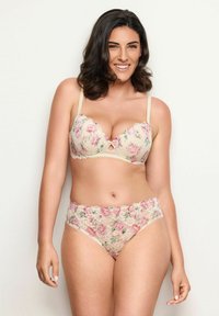 Žena s tmavými vlasy se usmívá, oblečená v korespondovaných květinových krajkových podprsenkách a kalhotkách v jemných béžových a růžových tónech na jednobarevném pozadí.