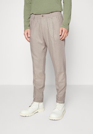 Homme portant un pantalon plissé beige, des bottes blanches à lacets et une chemise à manches longues vert clair, debout devant un fond uni.