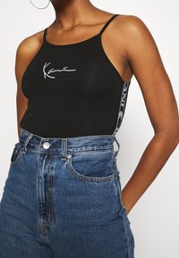 Ingrandimento di una donna che indossa un top nero con spalline sottili con la scritta "Karri" sul davanti e jeans blu a vita alta, con una mano in tasca.