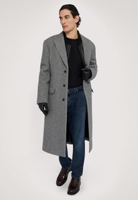 Grå ullrock med stora slag, svarta läderdetaljer, svarta handskar, mörka jeans och bruna läderskor. Modellen står mot en neutral bakgrund.