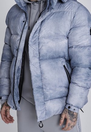 Homme portant une veste matelassée gris clair et un sweat à capuche gris assorti, les mains tatouées visibles, debout devant un fond uni.