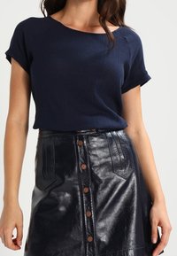 Marinblå stickad topp med rullade korta ärmar, matchad med en högmidjad kjol i svart konstläder som har bruna knappar och fickor framtill.